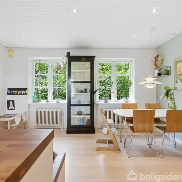 Et spisebord med træstole står i et lyst, moderne rum med store vinduer og udsigt til grønne omgivelser. Et sort vitrineskab står mellem vinduerne; en skål med frugt ses i forgrunden.