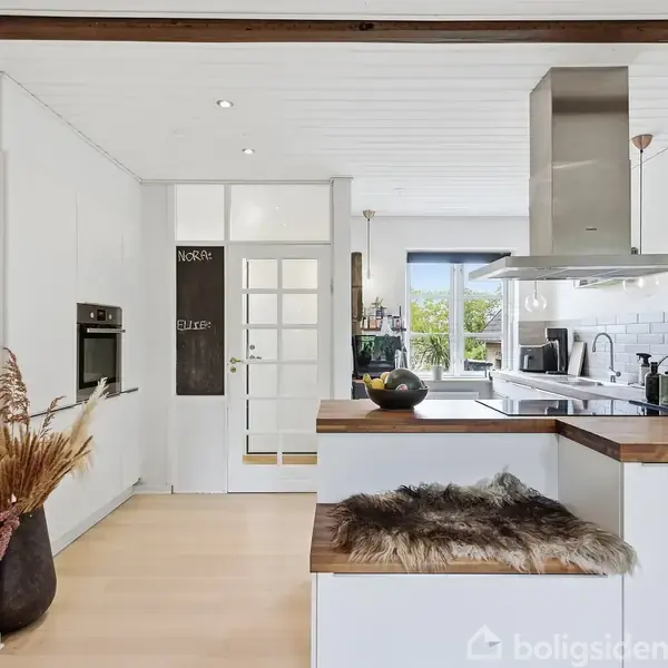 Køkken med hvidt interiør, træbordplade og emhætte. Dekoreret med planter og tørrede blomster. Låger er åbne til lys stue. Tekst på dør: "NORGE" og "SUITE".
