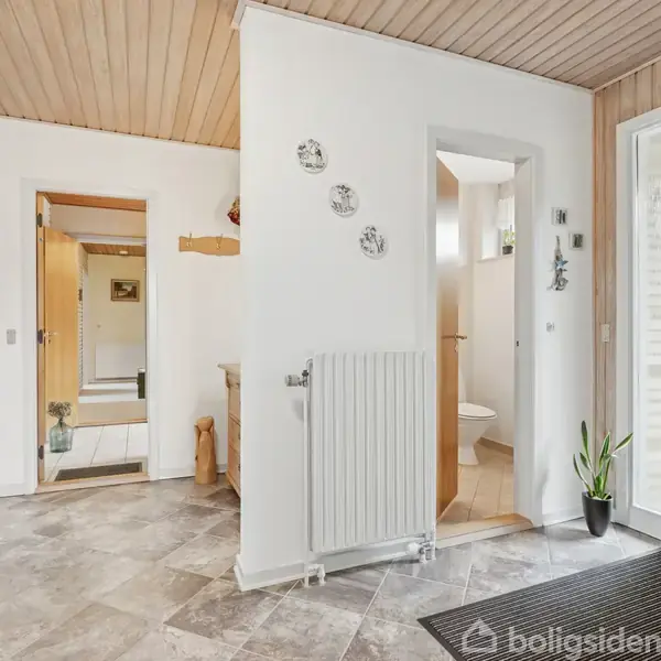 En entre med grå fliser, hvidmalede vægge og træloft. Der er en radiator, flere døråbninger til tilstødende rum, en potteplante ved en glasdør og vægdekorationer.