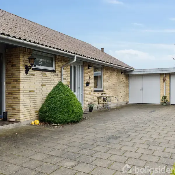 Et enfamiliehus med murstensfacade og fladt tag står ved siden af en stor garage. En sti af fliser fører til hoveddøren, omgivet af buske og udendørsmøbler.