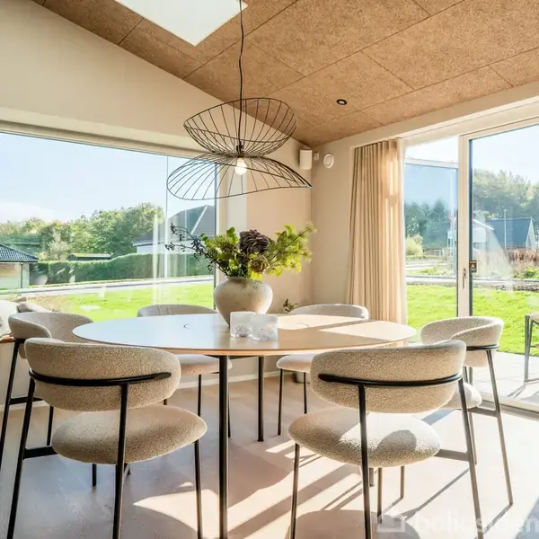 Et rundt spisebord med seks stole står i et lyst rum med store vinduer. En moderne lampe hænger over bordet, og udsigten viser en frodig have udenfor.