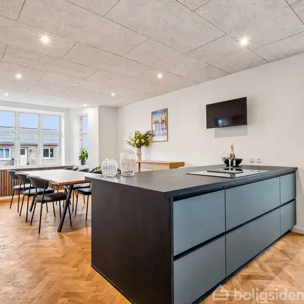 En moderne køkkenø med sorte skabe står centralt i et lyst rum. I baggrunden er et spisebord med stole, samt vinduer og en dør giver udsigt til udendørs træer.