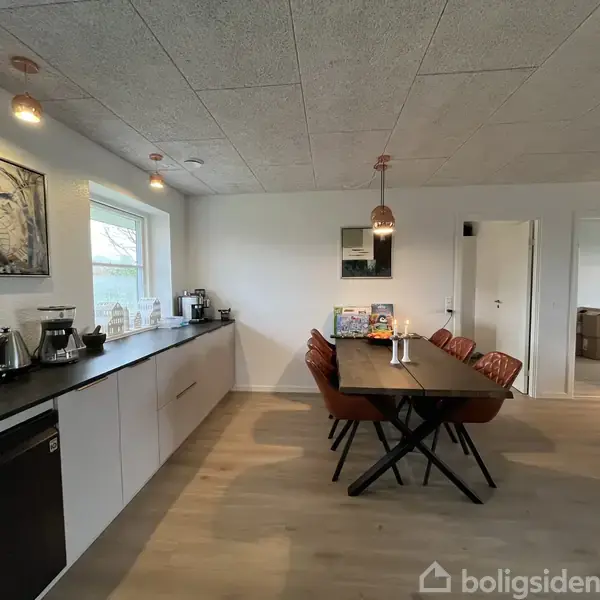 Et langt spisebord med seks brune stole står centralt i et lyst køkkenalrum. På køkkenbordet til venstre er der forskellige køkkenmaskiner og pyntegenstande ved et vindue.