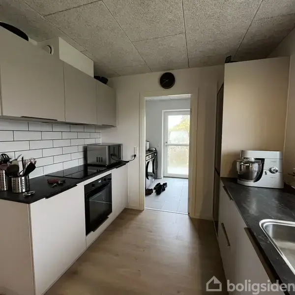 Køkken med hvide skabe og sorte bordplader. Kogeplade og ovn er til venstre. Der står køkkenredskaber og en røremaskine. En dør fører til et andet rum i baggrunden.