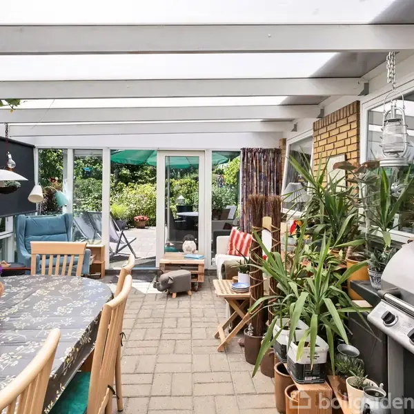 Overdækket terrasse med spisebord og planter. Bordet har en blomsterdug og omgives af træstole. Grill står ved siden af. I baggrunden ses en sofa og udsigt til en have.
