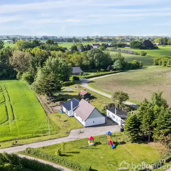 Hus uden handlinger omgivet af åbne marker og grønne træer. Der er også en legeplads med skydelåg og gynger i forgrunden.