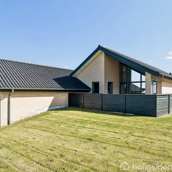 Et moderne hus med store vinduer ligger på en velplejet græsplæne. Huset har et skråt tag og lyse mursten, omgivet af en minimalistisk have under en blå himmel.