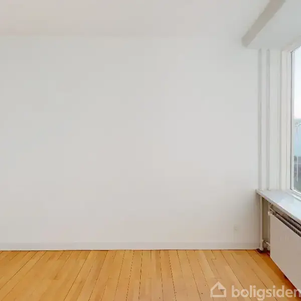 Et tomt rum med hvide vægge og et trægulv. Et stort vindue til højre giver udsigt til en græsplæne og en bygning udenfor.