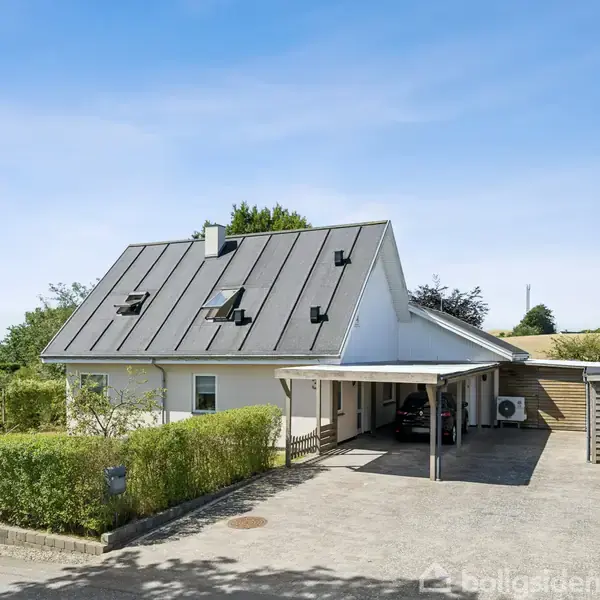 Hvidt hus med skråt tag og flere tagvinduer, står stille ved en flisebelagt indkørsel med carport. Omgivet af hække og grønne træer under en blå himmel.