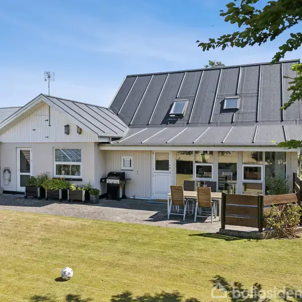 En hvid villa med mørkt tag står i et velplejet haveområde. En terrasse med havemøbler og grill er placeret ved siden af huset. Træer omkranser haven. En bold ligger på græsset.