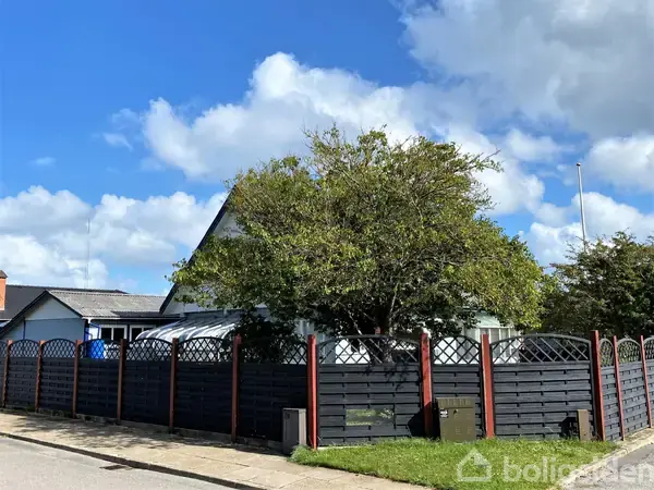 Et hus står bag et sort plankeværk med røde stolper. Et stort træ hænger over hegnet. Blå himmel og hvide skyer udgør baggrunden.