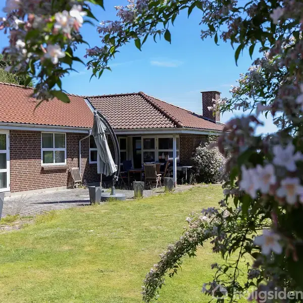 Murstenshus med rødt tegltag står i en grøn have. En parasol og havemøbler ses på terrassen. Forgrunden har blomstrende grene. Klar blå himmel i baggrunden.