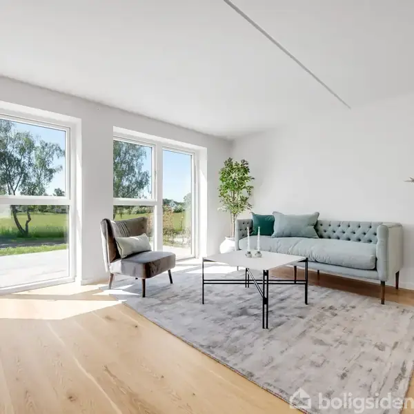 En stue med en grøn sofa og en brun lænestol placeret på et tæppe. Store vinduer lukker naturligt lys ind, og udenfor ses en græsplæne med træer.