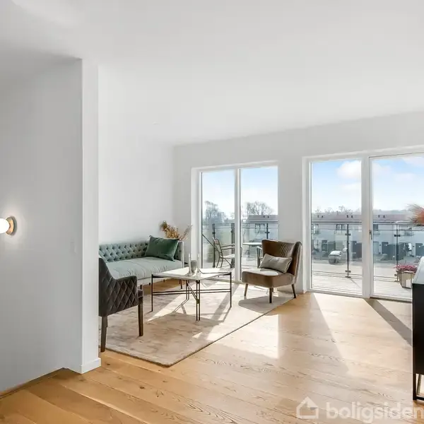 Stue med moderne møbler, inklusive en sofa og stole, står på et trægulv. Sollys strømmer ind gennem store vinduer, der fører ud til en altan med udsigt over bygninger.