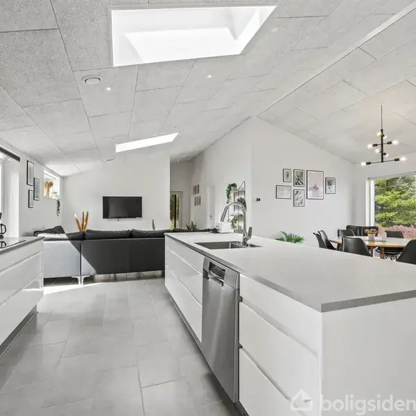 En moderne køkkenø står centralt, med indbyggede hvidevarer og opvaskemaskine. Omkring er en åben stue med sofagruppe, fladskærms-TV og et spisebord, badet i naturligt lys fra ovenlysvinduer.