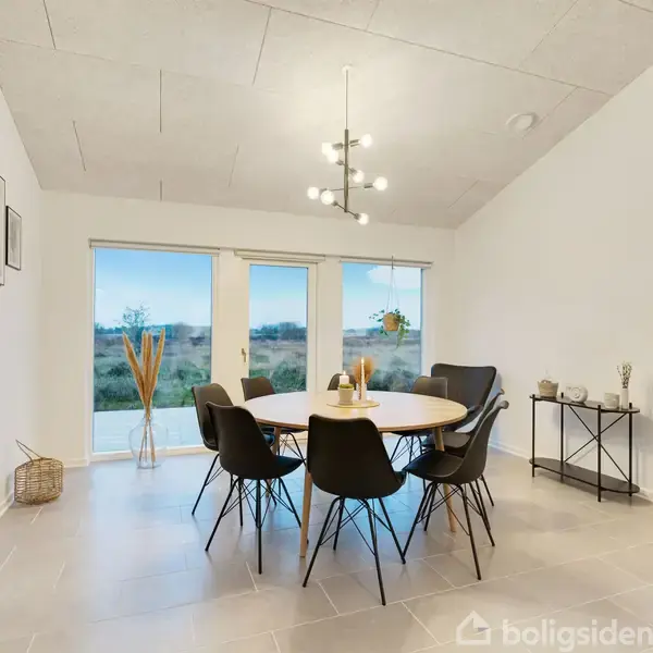 Et rundt spisebord med seks stole står i et lyst rum med store vinduer, der viser en udsigt over landskabet. Rummet er dekoreret med vægbilleder og planter.