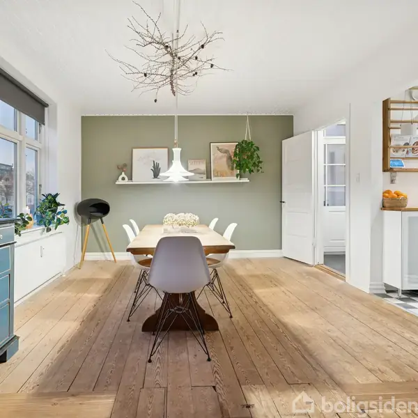 Spisebord omgivet af stole står på trægulv i et lyst spisestue. Moderne lysekrone hænger over bordet. Grønne planter og et malet skab dekorerer rummet, som har adgang til køkkenet.