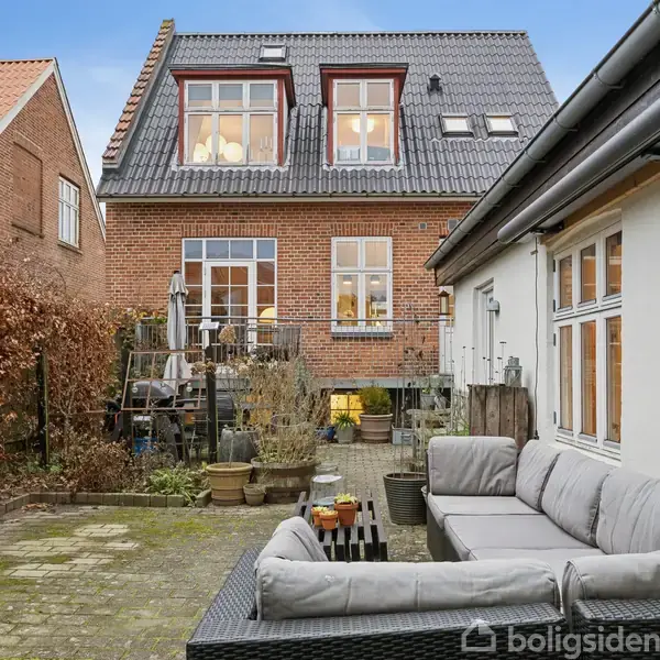 To-etagers murstenshus med mange vinduer og mørkt tag. Foran er en gårdhave med grå sofa, borde og planter, omgivende af en hæk og naboens husvæg.