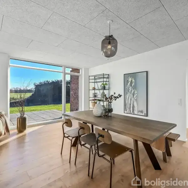 En spisestue med et langt træbord og stole ved siden af et stort vindue, der viser en have udenfor. En dekorativ vase står på bordet, og der hænger en lampe over det. På væggen ses et kunstværk, og der er en åben dør til et andet rum med dagslys.