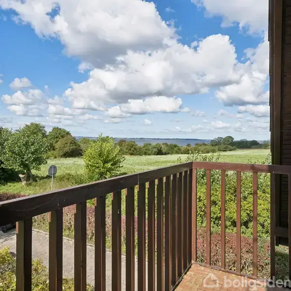 Balkon med trægulv og rækværk strækker sig ud fra en bygning. Udsigt over grønt landskab med træer og marker under en blå himmel med skyer.