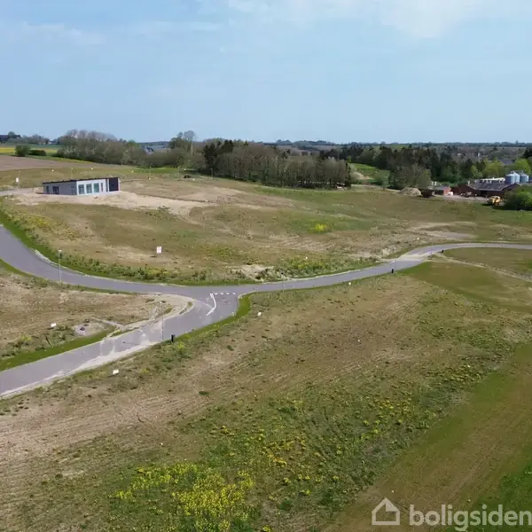 En snoet asfalteret vej løber gennem en åben mark med spredt græs og små buske. En ensomtliggende moderne bygning ses til venstre, omgivet af en landlig og let kuperet panorama.