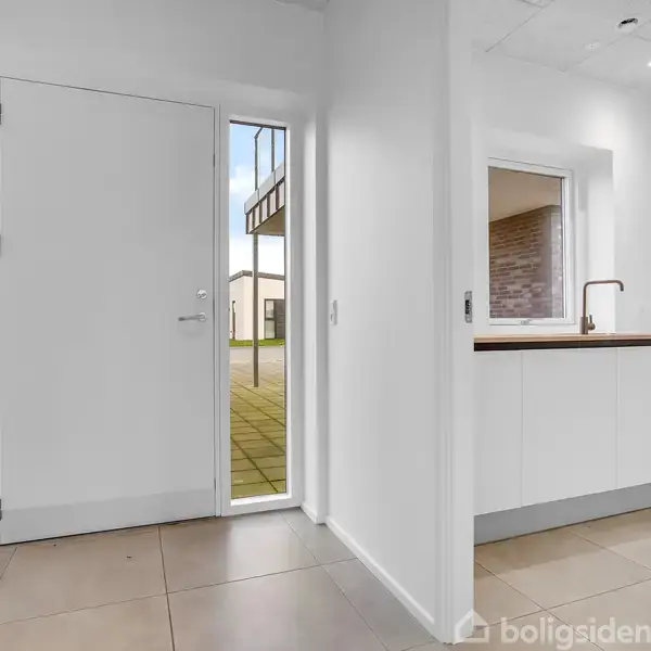 Hvid dør står åben mod en gårdhave, omgivet af fliser. Til højre er en køkkenåbning med hvidt skab og indbygget vask. Indenfor ses lyse flisegulve.