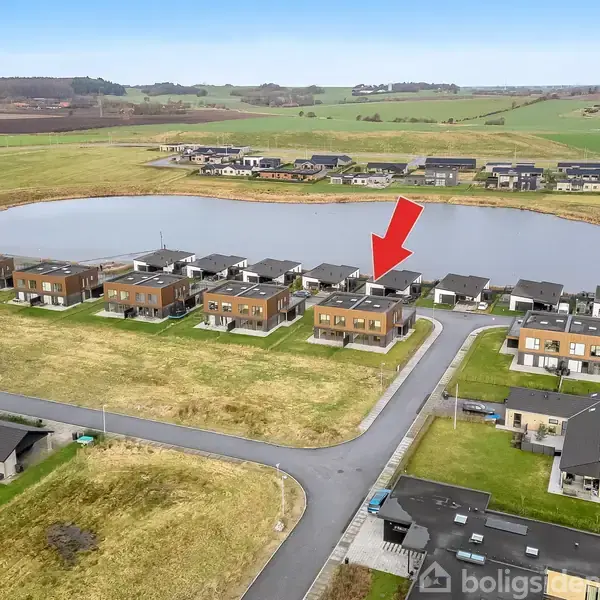 Rækkehuse står i en klynge ved en sø, omgivet af åbne marker. En rød pil peger på et specifikt hus.