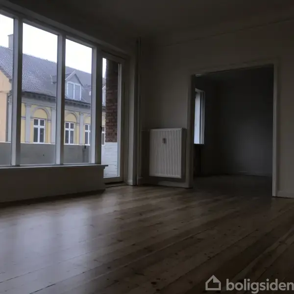 Et tomt rum med trægulv og store vinduer, der lader naturligt lys strømme ind. Udsigt til en gul bygning udenfor. Radiator placeret på væggen ved siden af en åbning til et andet rum.