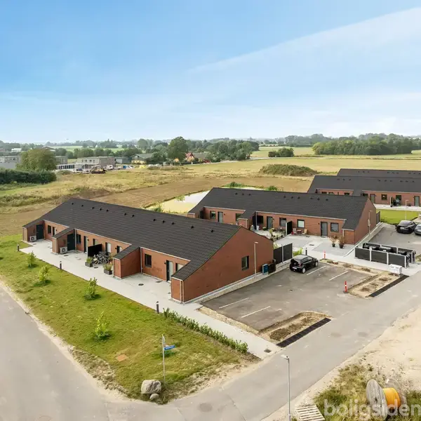 Rækkehuse med sorte tage og brune mursten står i et åbent, landligt område. Der er parkeringspladser foran, omgivet af græs og marker i baggrunden.