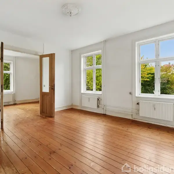 Et rummeligt, tomt værelse med trægulv, hvide vægge og to store vinduer, som giver udsigt til grønne træer udenfor. En åben dør fører til et tilstødende rum.