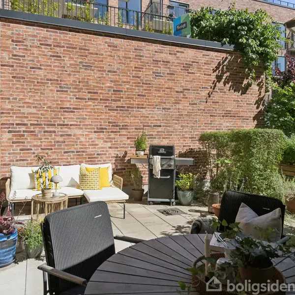 Terrassen har møbler som en sofa og et rundt bord med sorte stole. En grill ved siden af en murstensvæg. Grønne planter omkranser området, skaber en hyggelig atmosfære.