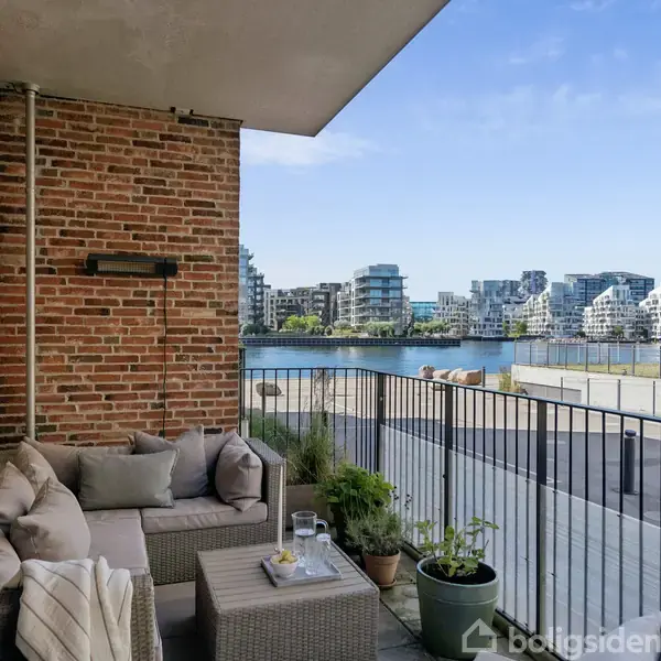 Grill står stille på terrasse omgivet af frodig have med krukker og planter. Omkranset af murede bygninger med altaner, hvilket skaber en hyggelig byhave-atmosfære.