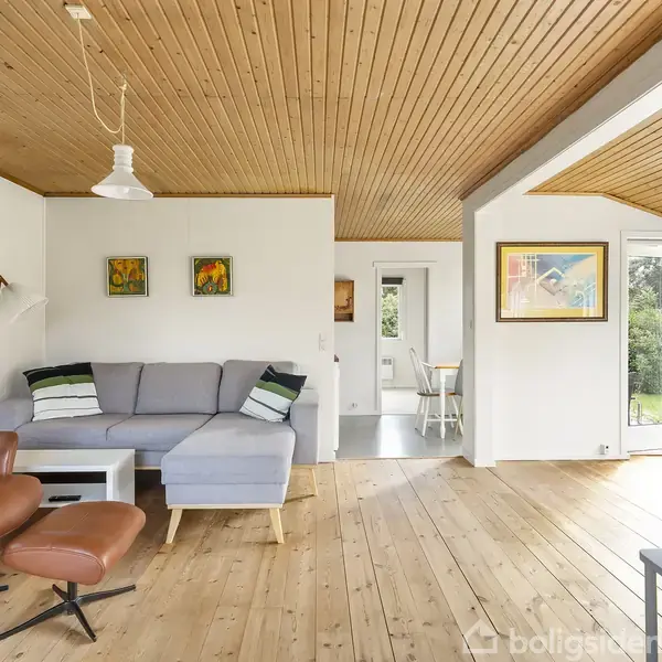 Stue med gråt hjørnesofa og brunt læderlænestol. Sofaen står på trægulv ved siden af hvide vægge dekoreret med kunst. Rummets store vinduer giver udsigt til en grøn have.