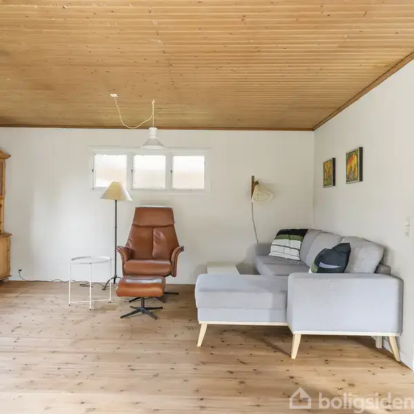 Stue med gråt hjørnesofa, brun lænestol og lyst træloft. Træskænk til venstre med bøger. To små vinduer over lænestolen, lyse træplanker på gulvet.