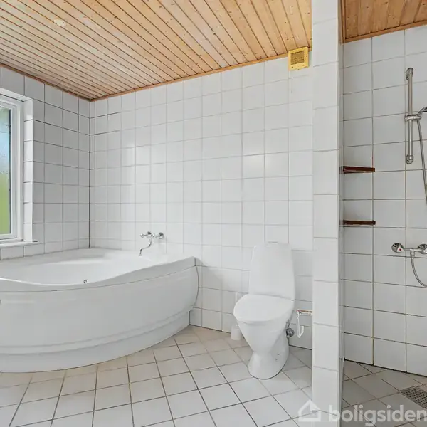 Et hjørnebadekar under et vindue, en toiletstol og en bruser i et flisebelagt badeværelse med træloft. Væggene og gulvet er dækket af hvide fliser.