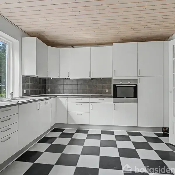 Et moderne køkken med hvide skabe og sorte fliser ser ud mod en have gennem store vinduer. Tjekkede sort-hvide gulvfliser giver et stilrent look.