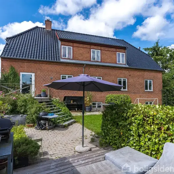 Rød murstensvilla med blåtag sidder i en have. En terrasse med parasol og planter er i forgrunden, omgivet af grønne buske og havemøbler under en klar blå himmel.