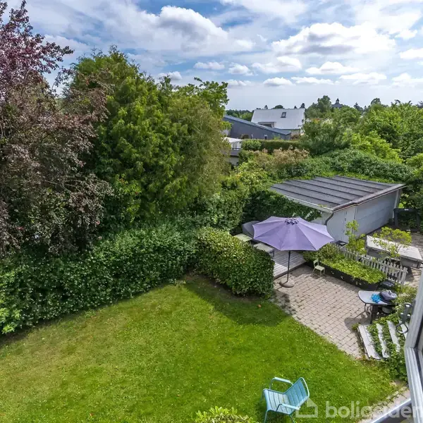 Grøn have med græsplæne og terrasse. På terrassen er der parasoller og stole. Omgivet af både tætte buske og høje træer, under en delvist skyet blå himmel.