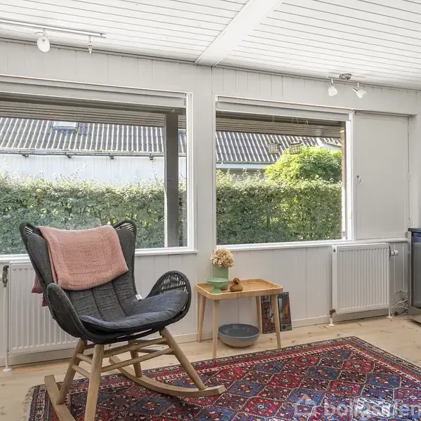 Gyngestol med lyserød pude står stille i lyst rum. Kirsebærsbord med blomstervase ved siden af. Tæppe dekorerer trægulvet, mens store vinduer viser hæk udenfor.