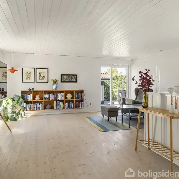 Stue med lyst trægulv og hvidt loft, møbleret med reol fyldt med bøger, et lille sofabord og lænestole i hjørnet. Store vinduer giver lys. En vase med blomster pryder et bord.