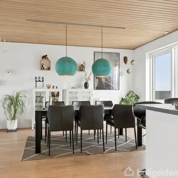 Spisebord med otte sorte stole står under to grønne lamper i et lyst rum med trægulv og hvid reol. Vægge prydes af kunstværker og grønne planter.
