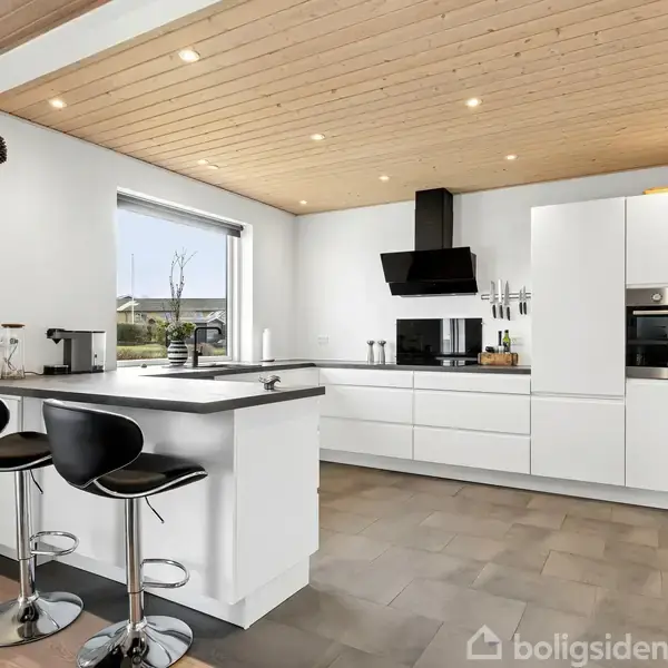 Et moderne køkken med hvide skabe og en ø-ø med to sorte barstole. Vinduet giver udsigt til en have, mens en træbeklædt loft og indbyggede spots skaber lys atmosfære.