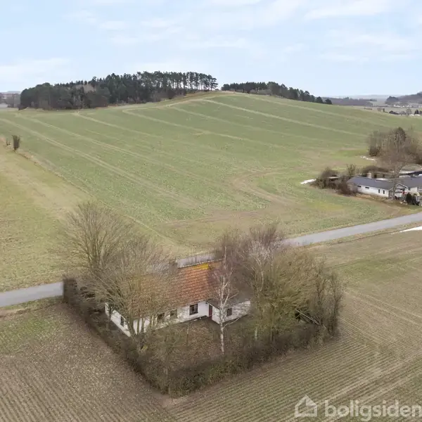 Et hus ligger omgivet af træer og marker. I baggrunden ses en landevej og flere marker med spredte træer og en række huse. Himmel er overskyet.