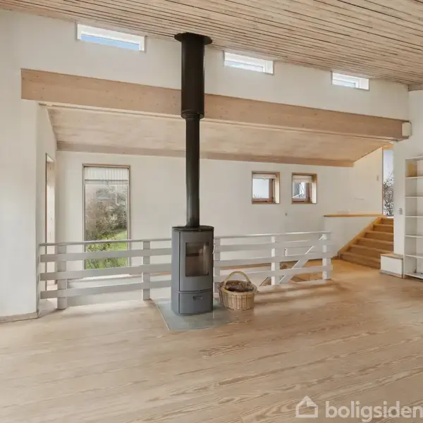 Brændeovn står i midten af et lyst stueområde med trægulv og -loft. Til venstre er store vinduer og en dør, mens til højre ses en trappe og reoler.