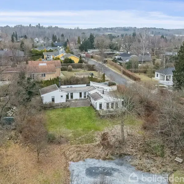 Et hvidt hus med fladt tag ligger i en villakvarter, omgivet af træer og græs. En vej løber tæt ved, og en lille sø ses forrest i billedet.