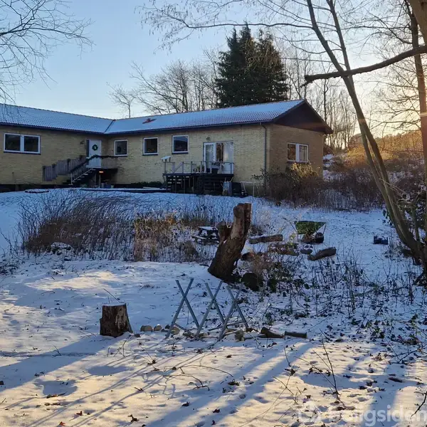 Et gult, lavt hus med trappe i midten står i et sneklædt landskab med træer og buske. En bil er parkeret til venstre, og solen skinner gennem grenene.