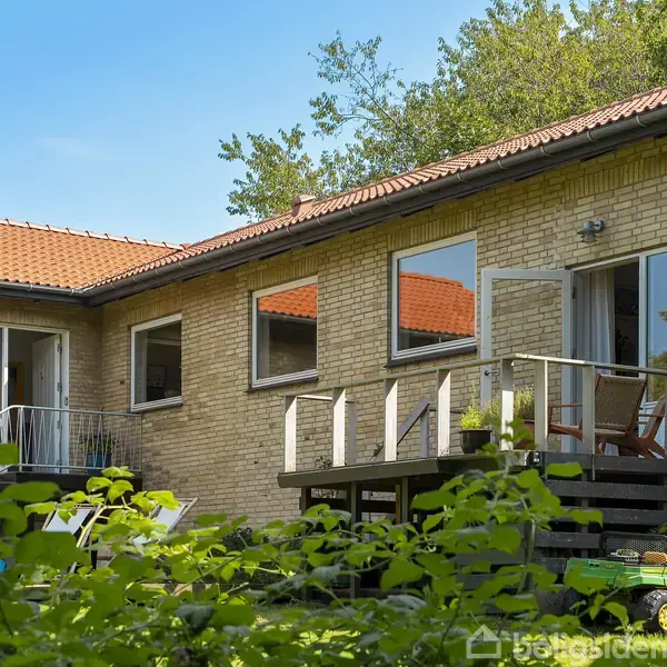 Et én-etagers hus med murstensvægge og rødt tegltag, står i en frodig have. Træterrasse med stole er synlig, omgivet af tæt vegetation og træer i baggrunden.