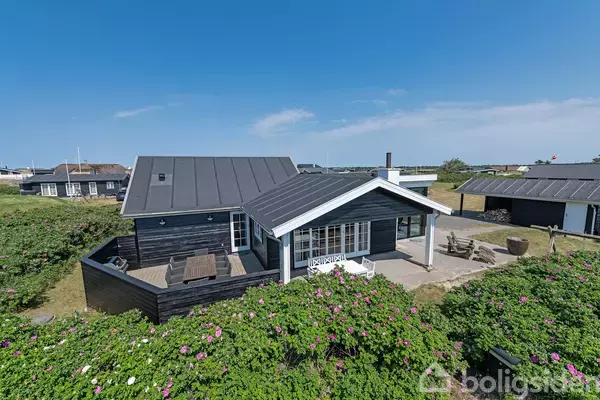 Sort træhus med terrasse står stille i en sommerhusområde omgivet af grønne buske og blomster. Klar blå himmel ovenover skaber en rolig atmosfære.