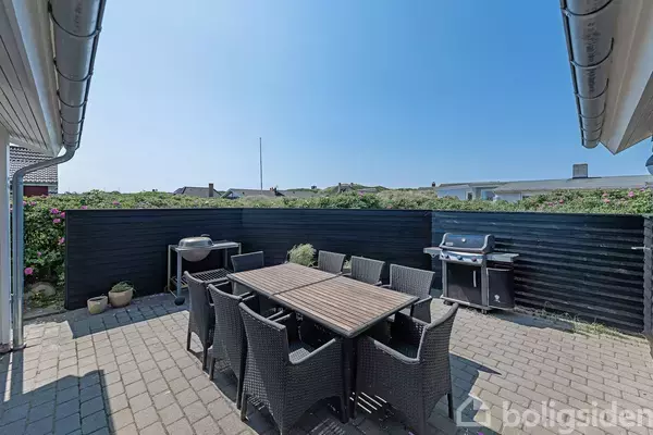 Udendørs terrasse med havebord omgivet af ni flettede stole. To grillapparater står mod et sort plankeværk. Omgivelserne er solrige, med en blå himmel overhead.