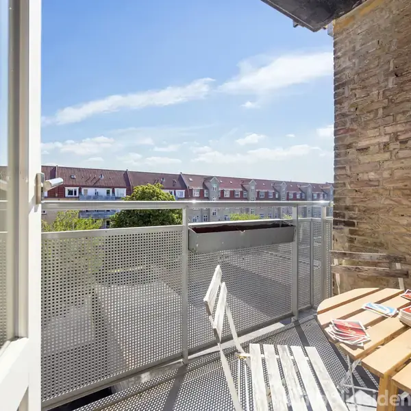 En balkon med et lille rundt bord og to stole står udendørs. Bordet har en flaske vand og et glas på. Baggrunden viser rækkehuse under en blå himmel med spredte skyer.